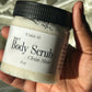 Hand holding a jar of 'Body Scrub' with a brush on a light fabric background