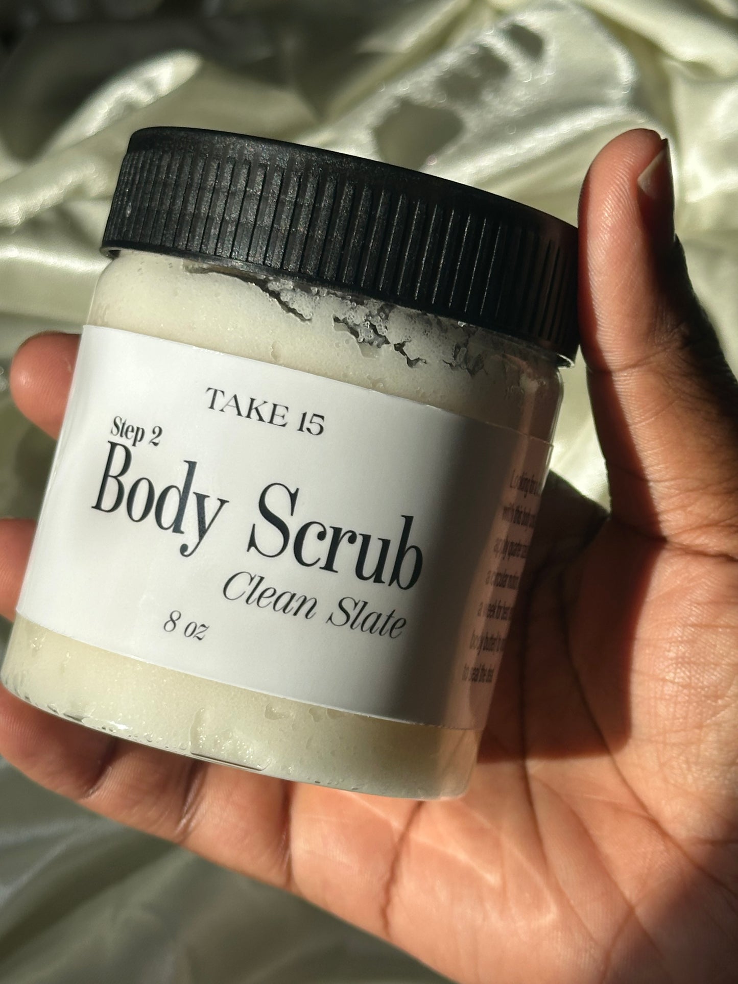 Hand holding a jar of 'Body Scrub' with a brush on a light fabric background